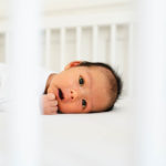 Neugeborenes Baby im Babybett schaut in dei Kamera durch die Stangen des Bettes