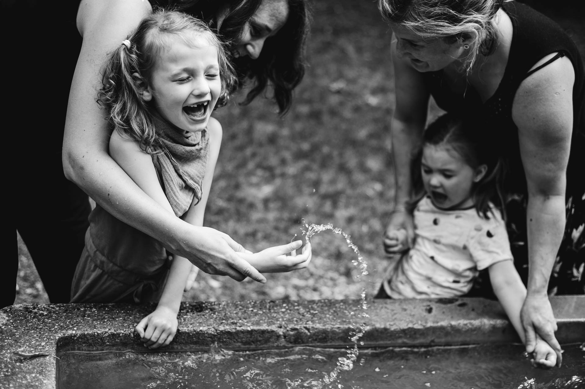 Zwei Mädchen mit Rett Syndrom und ihre Mütter spielen mit Wasser an einem Brunnen im Freien
