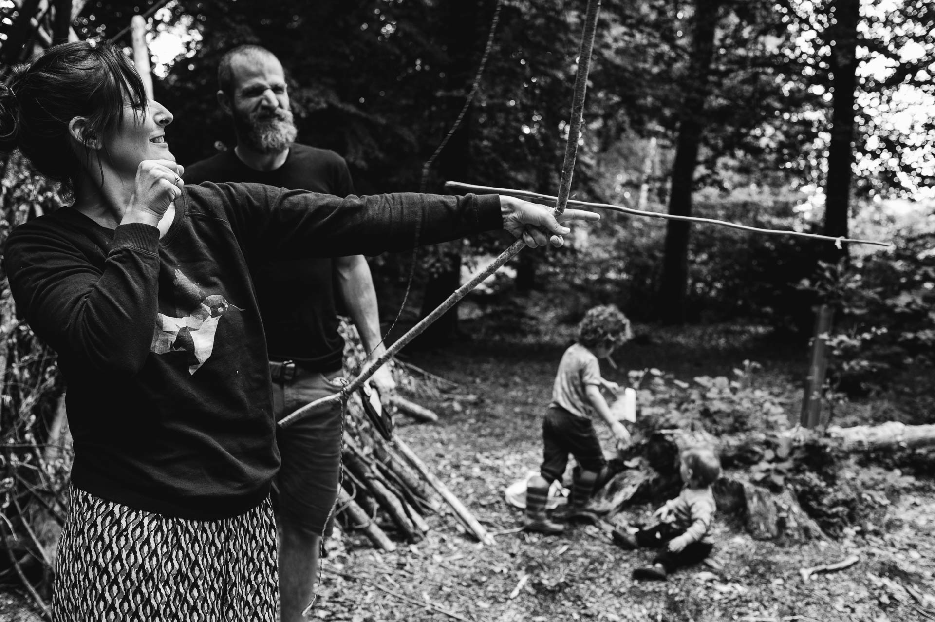 Frau wirft einen Ast mit Pfeilbogen im Wald und Mann schaut zu während die Kinder im Hintergrund spielen