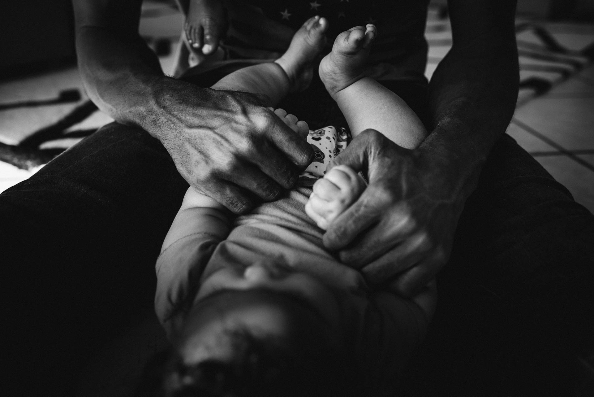 Detailbild von den Händen eines Vaters, die die Hände seines Babys hält, ein Fuss des älteren Kindes, das auf den Schultern des Vaters sitzt, ist sichtbar